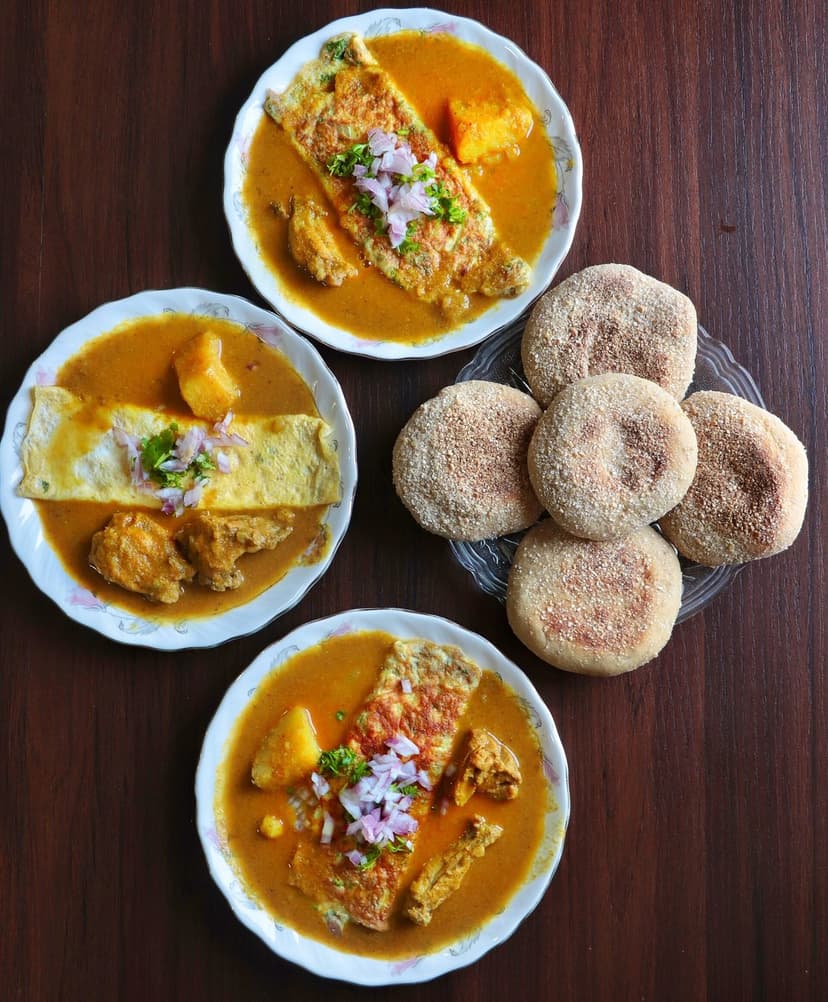 Three plates of Ross omelette, an omelette in spicy orange meat gravy topped with onion, beside a basket of round Goan poi bread