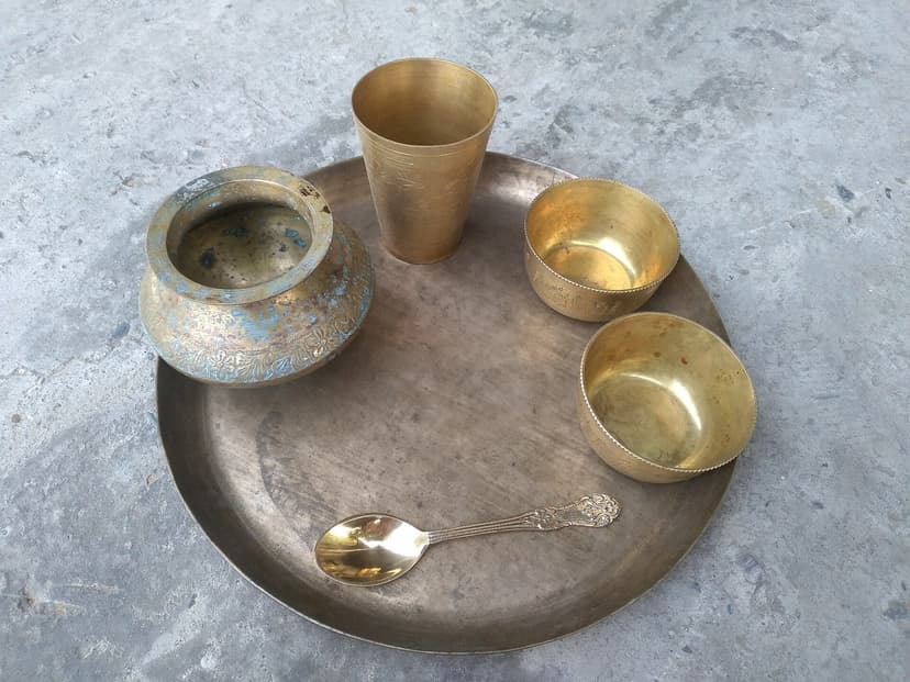 A traditional Indian brass thali set on a stone surface — large round paraat, drinking lota, etched glass, two katoris and an ornate spoon, all in well-aged brass that Boti families and Himachali households use for ceremonial meals