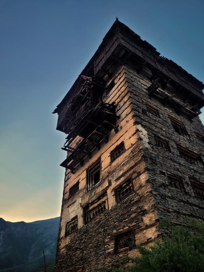 Medieval Gondhla Fort in Lahaul, a seven-to-eight-story tower of stone and timber with projecting carved wooden balconies, photographed from below at dusk