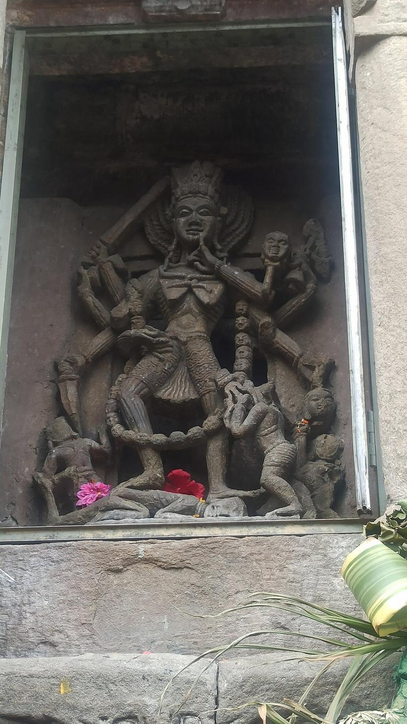 Ancient stone relief sculpture of a multi-armed deity set in a niche on the outer wall of Baijnath Temple with fresh flowers left by devotees