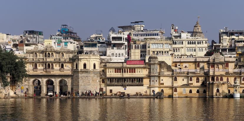 The cream-coloured havelis and stone steps of Gangaur Ghat and Bagore Ki Haveli rising straight from Lake Pichola in Udaipur, with people at the water edge and reflections in the lake
