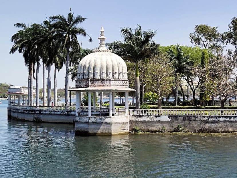 A white domed chhatri pavilion on the edge of Fateh Sagar Lake in Udaipur, framed by tall palm trees under a clear blue sky.
