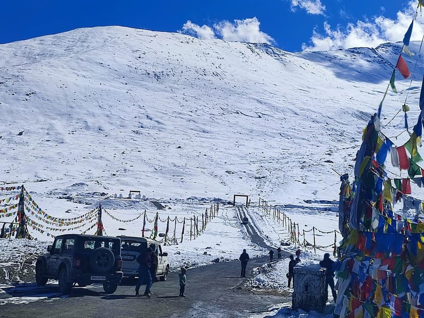Kunzum La at 4,551 m on a snow day, chain-and-rope barrier across the highest point, prayer flags on the right, a black SUV waiting on the cleared snow road, the shoulder of the pass white above