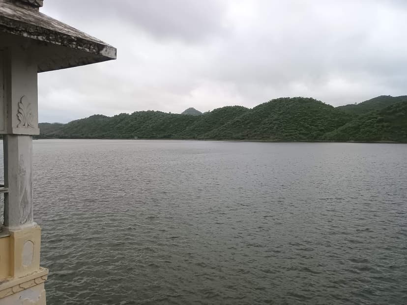Calm grey water of Badi lake ringed by green Aravalli hills under a cloudy sky, a stone chhatri pavilion at the left edge