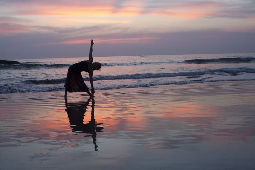 A person in triangle pose silhouetted on a wet beach at sunset, the pink and orange sky reflected in the sand