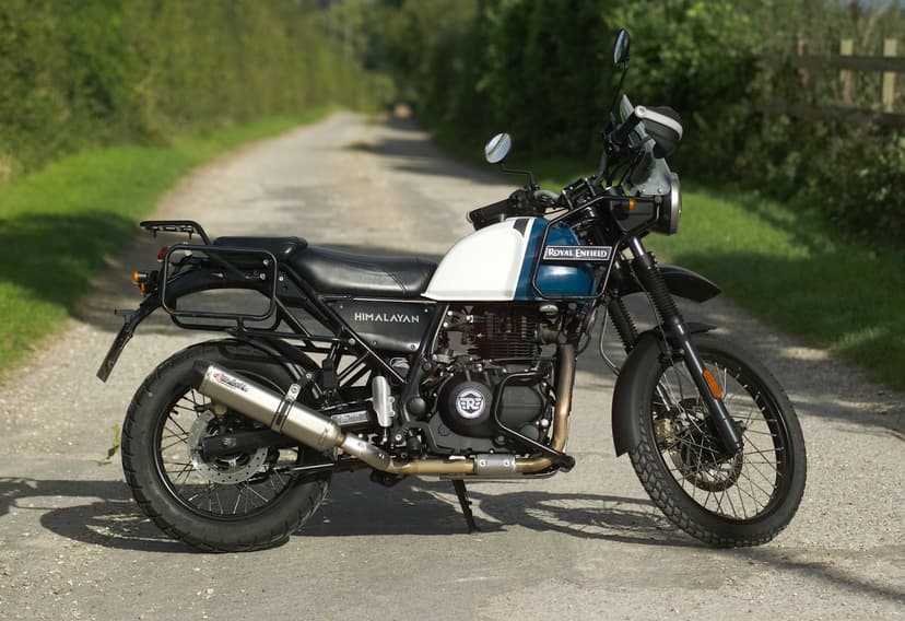 Side profile of a Royal Enfield Himalayan adventure motorcycle with white and blue tank, long-travel front suspension, exposed tubular frame and high-set exhaust