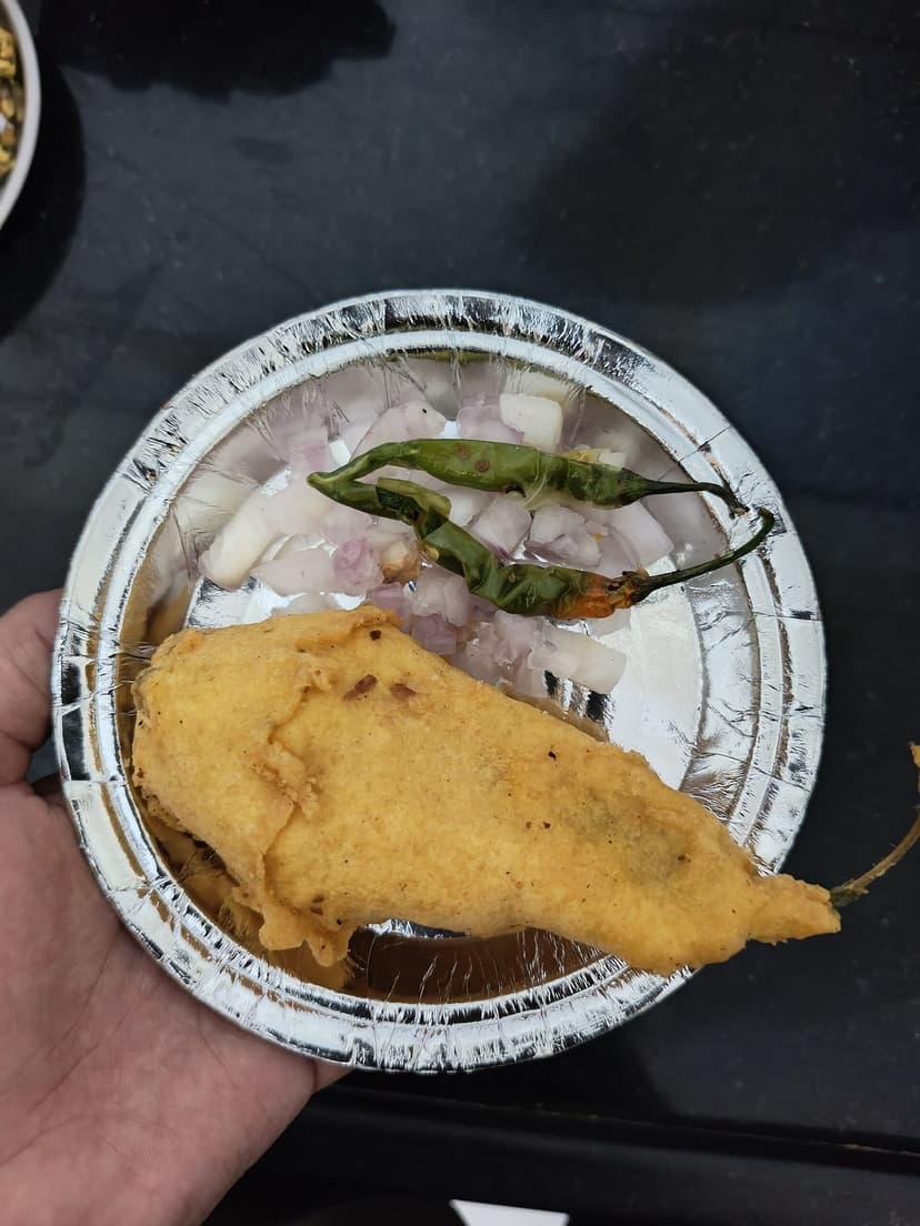 A mirchi bada, a whole green chilli in gram-flour batter, deep-fried, on a foil plate with raw onion and green chillies on the side, held in a hand