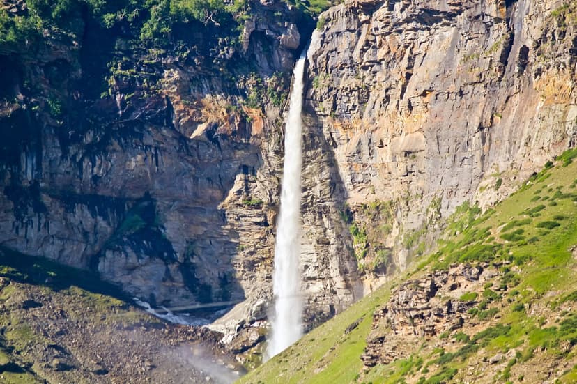 Tall single-drop Sissu waterfall cascading down a sheer brown-grey cliff with a green grassy slope in the foreground, Lahaul valley