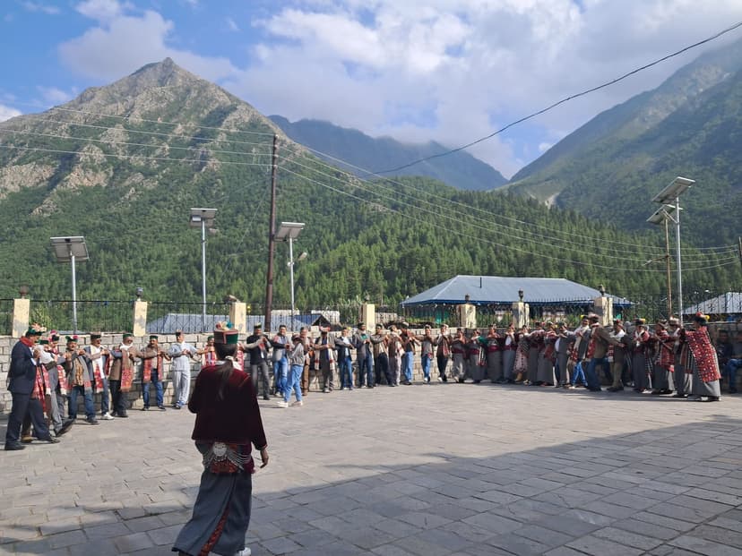 Long line of Kinnauri dancers in maroon-and-grey traditional dress with red caps performing in a paved village square at Chitkul with Baspa valley peaks rising behind