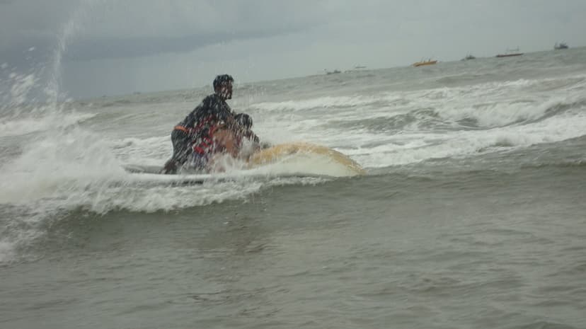 A man in a life jacket riding a jet ski with a pillion through choppy grey surf, fishing boats dotted on the horizon under an overcast sky