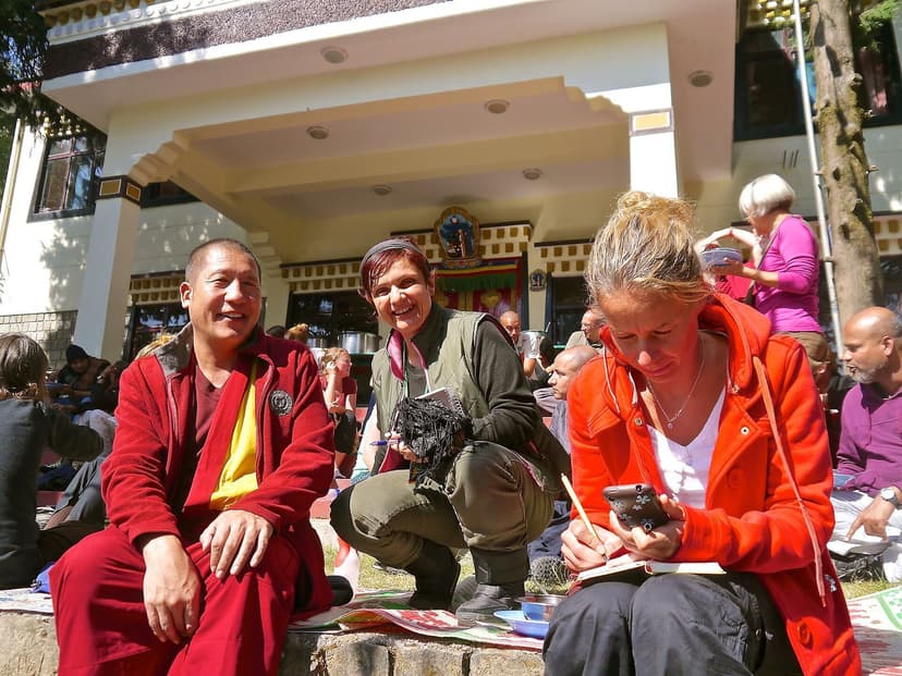 A Tibetan monk in maroon robes and a visitor writing notes among a seated group outside a temple in McLeod Ganj