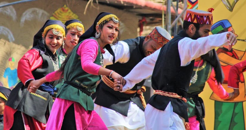 Mixed-gender Nati troupe mid-step with women in pink kurtas, green velvet sadri vests, silver dhattu head ornaments, men in white tunics and embroidered Kulluvi topi, all linked in a criss-cross hand pattern