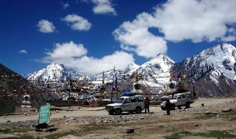 Kunzum Pass at 4,551 metres between Lahaul and Spiti valleys, with prayer flags, parked 4x4 vehicles, and snow-capped peaks under blue sky