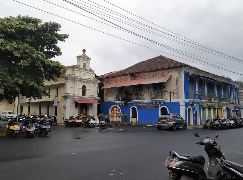 A street in the São Tomé quarter of Panjim on a rainy day, with a small white chapel, a blue Portuguese-era building, parked scooters and overhead wires.
