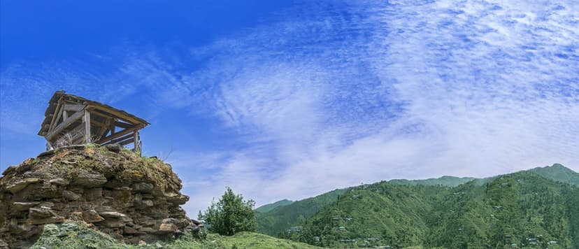 Panoramic view of Janjehli Valley in Seraj region, Mandi district, with a stone ruin on a ridge in the foreground and forested slopes with scattered settlements on the far side
