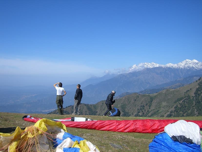 Three people standing on a grassy ridge with paraglider canopies in red blue and yellow spread out behind them, snow-capped Dhauladhar peaks in the distance