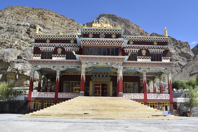 The painted facade of Sakya Tangyud Monastery in Kaza. Colourful red, gold and blue Tibetan motifs across the wood-and-stone front, central staircase leading up to the entrance, brown cliff face rising directly behind