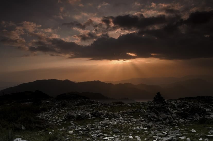 Evening sunset from a high ridge in the Great Himalayan National Park near Shoja, dark clouds breaking over silhouetted Himalayan mountain layers, golden rays filtering through onto grass and scattered rocks in the foreground