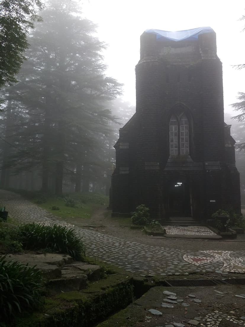 The dark basalt neo-gothic facade of St John in the Wilderness church between Forsyth Ganj and McLeod Ganj, tall deodars in morning mist, cobbled stone path curving up to the arched entrance, moss on the lower stones