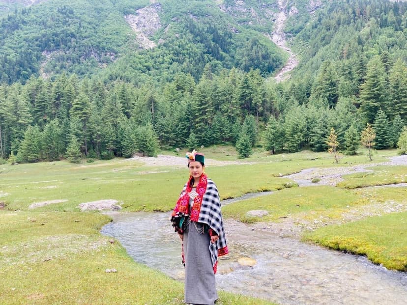 A woman in traditional Kinnauri dress with a red-bordered patterned shawl and flowered cap by a stream in a green Kinnaur meadow