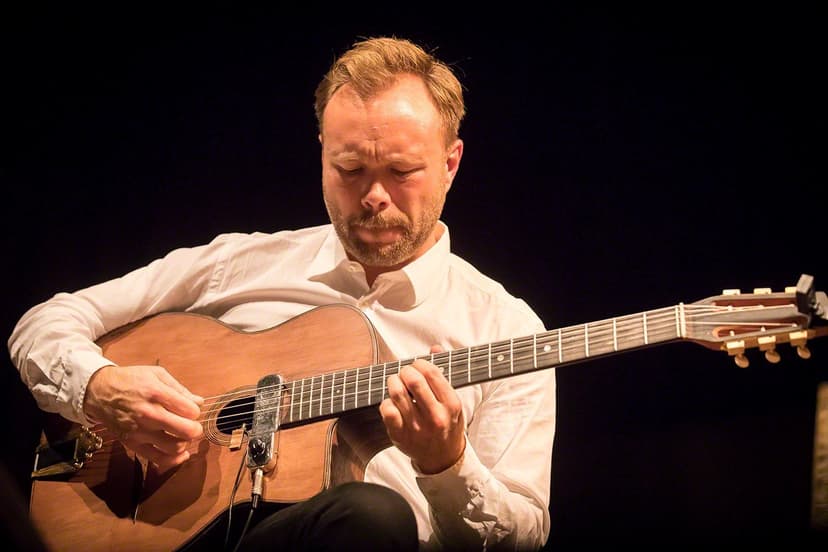 A close warm portrait of a man playing an acoustic guitar against a dark background, eyes down, absorbed in the music