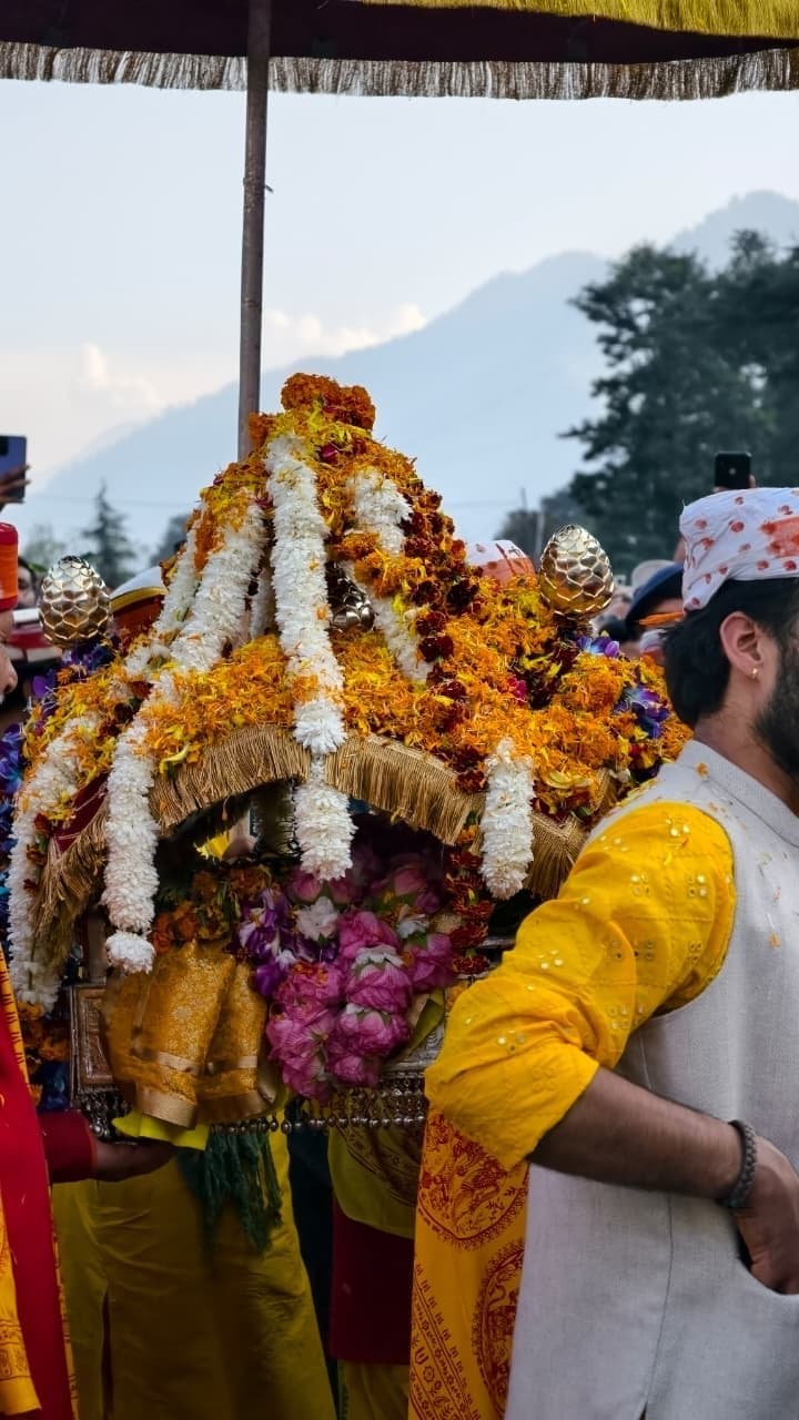 A Kullu deity palanquin shot from below at Dussehra 2025, silver kalash finials on top, fringed gold canopy, garlands of orange marigold and pink rose, a young devotee in a yellow kurta with red-printed gamcha standing beside it