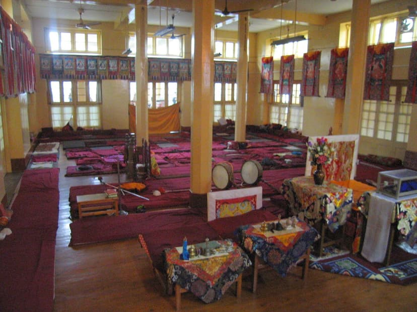 Teaching hall at the Dalai Lama Temple in McLeod Ganj showing ground-level seating with red cushions, drums, and thangka paintings