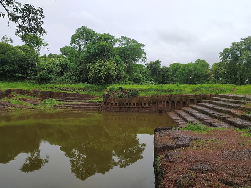 An old laterite-stone stepped temple tank with niches and steps down to still green water, ringed by monsoon greenery, on Divar Island