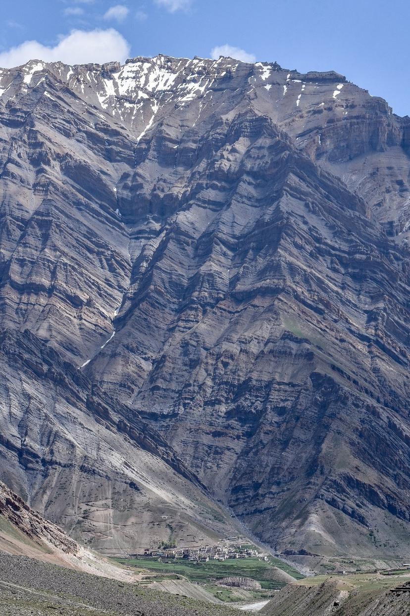Mud village under towering vertical cliffs in Pin Valley, with a small green field in the foreground