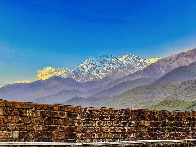 Snow-capped Dhauladhar peaks rising above the foothills, framed by a stone wall in the Baijnath Vaidyanatha temple complex
