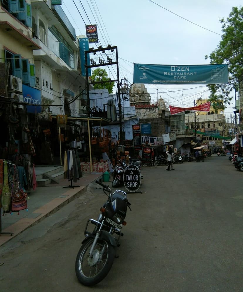 A working street in the old city of Udaipur with a tailor shop, a leather goods shop, a parked motorbike, power lines, a cafe banner strung across the lane and a temple spire behind