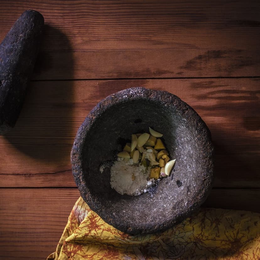 A stone mortar and pestle holding garlic, ginger, salt and spices ready to be ground into a fresh masala, on a wooden surface