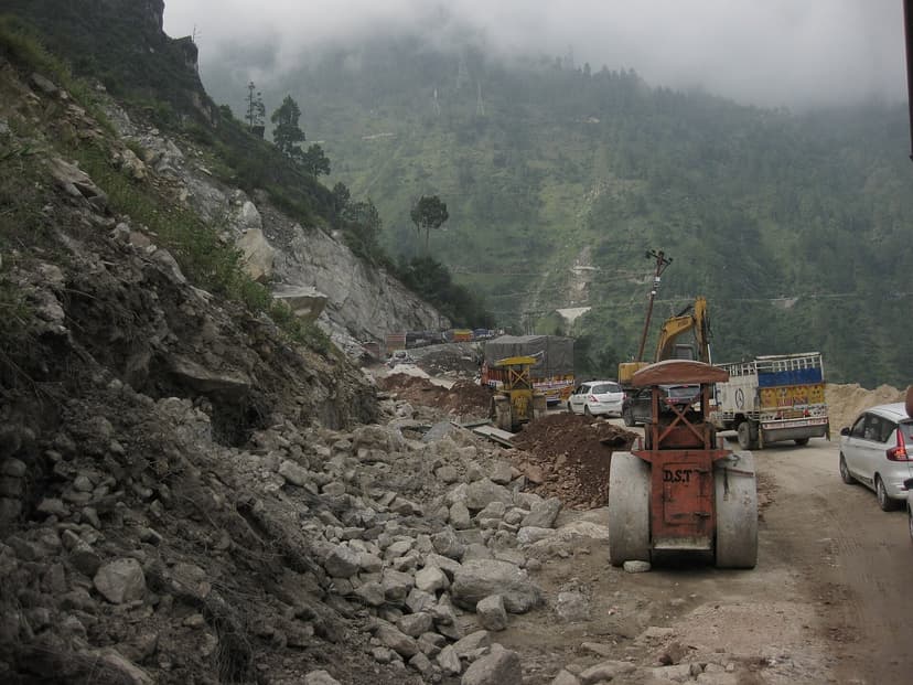 NH-5 in Kinnaur blocked by a landslide at Nigulsari with a road-roller clearing debris while trucks wait, misty hills behind