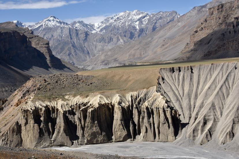 Eroded grey mud cliff formations with snow-capped Himalayan peaks and a dry brown valley behind, classic Spiti cold-desert landscape