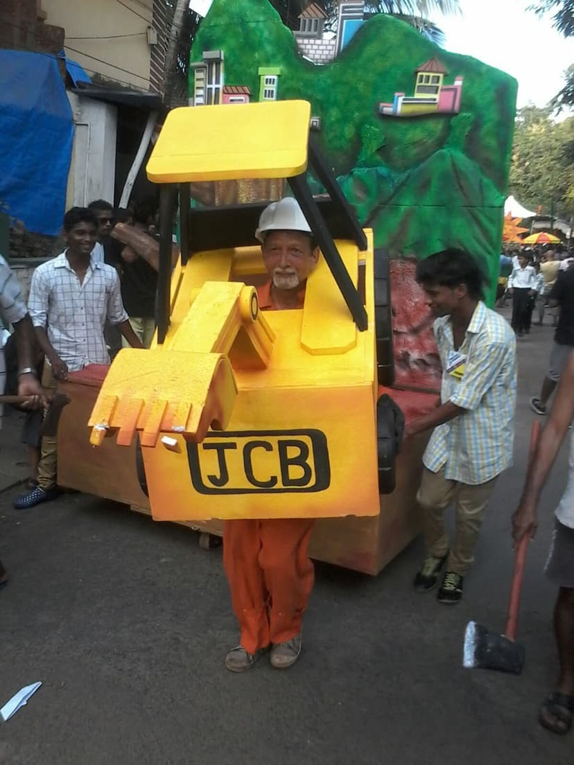 A man in a hand-made yellow JCB-excavator costume in a Bonderam street parade on Divar, a float of a green hillside with colourful houses behind him