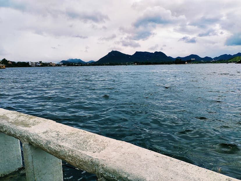 Fateh Sagar lake under a cloudy monsoon sky with green Aravalli hills across the water and a stone railing in the foreground