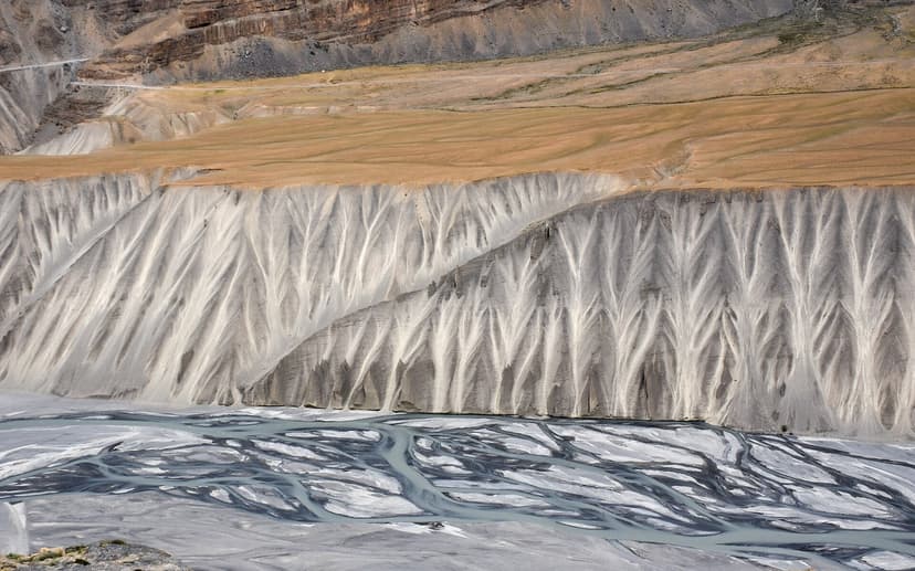 The eroded grey-white badlands above the Spiti river along NH-505 between Kaza and Losar, a flat brown table of farmland sitting on a wall of vertical erosion stripes, the river braiding below in pale blue channels