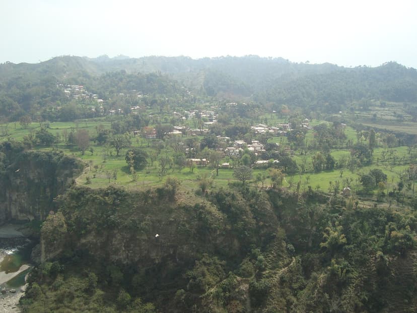 A wide aerial view of Kangra valley from Kangra Fort — green farms, scattered village houses, low forested ridges and hazy hills in the distance, the broad rural floor where Boti chef families like the one at Pathiar live and cook