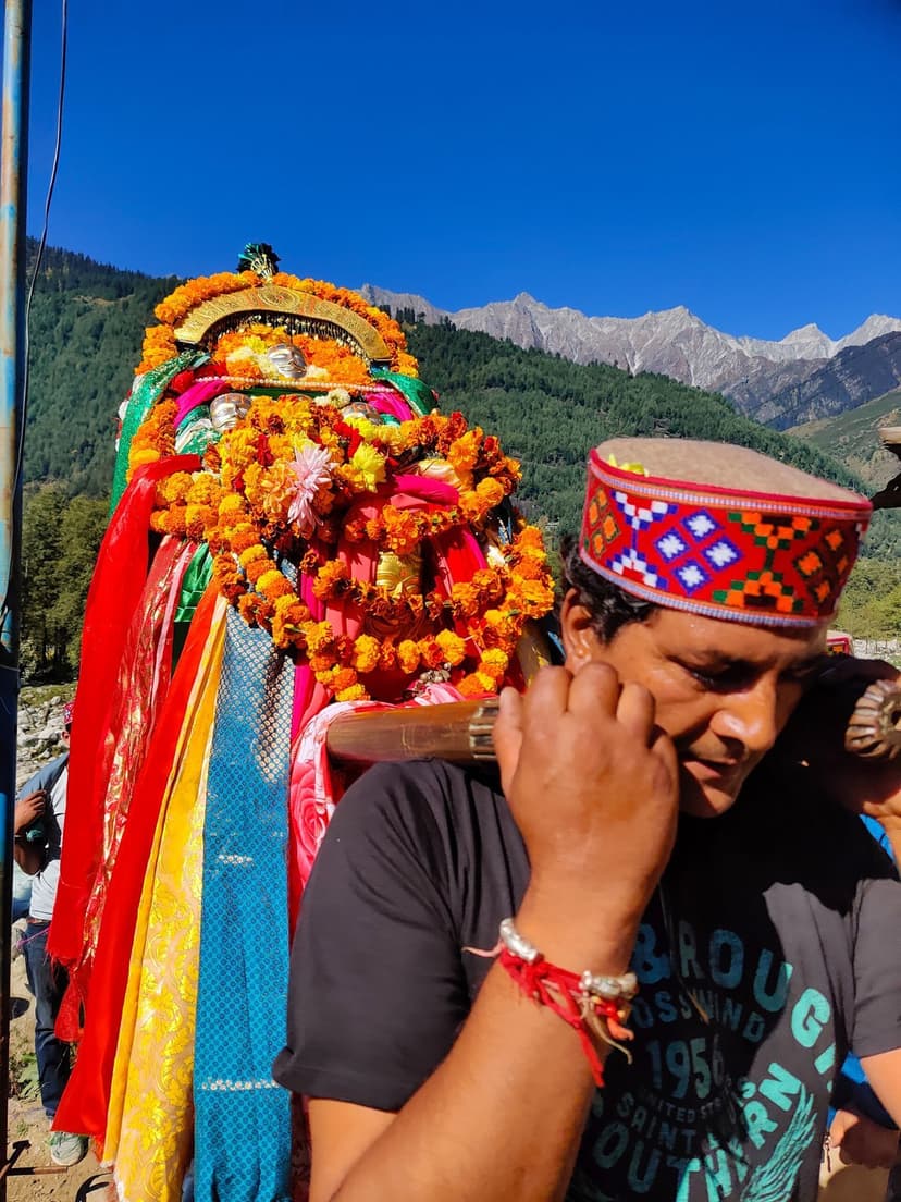 A man in a Kullu cap carries a garlanded deity palanquin on his shoulder with mountains behind