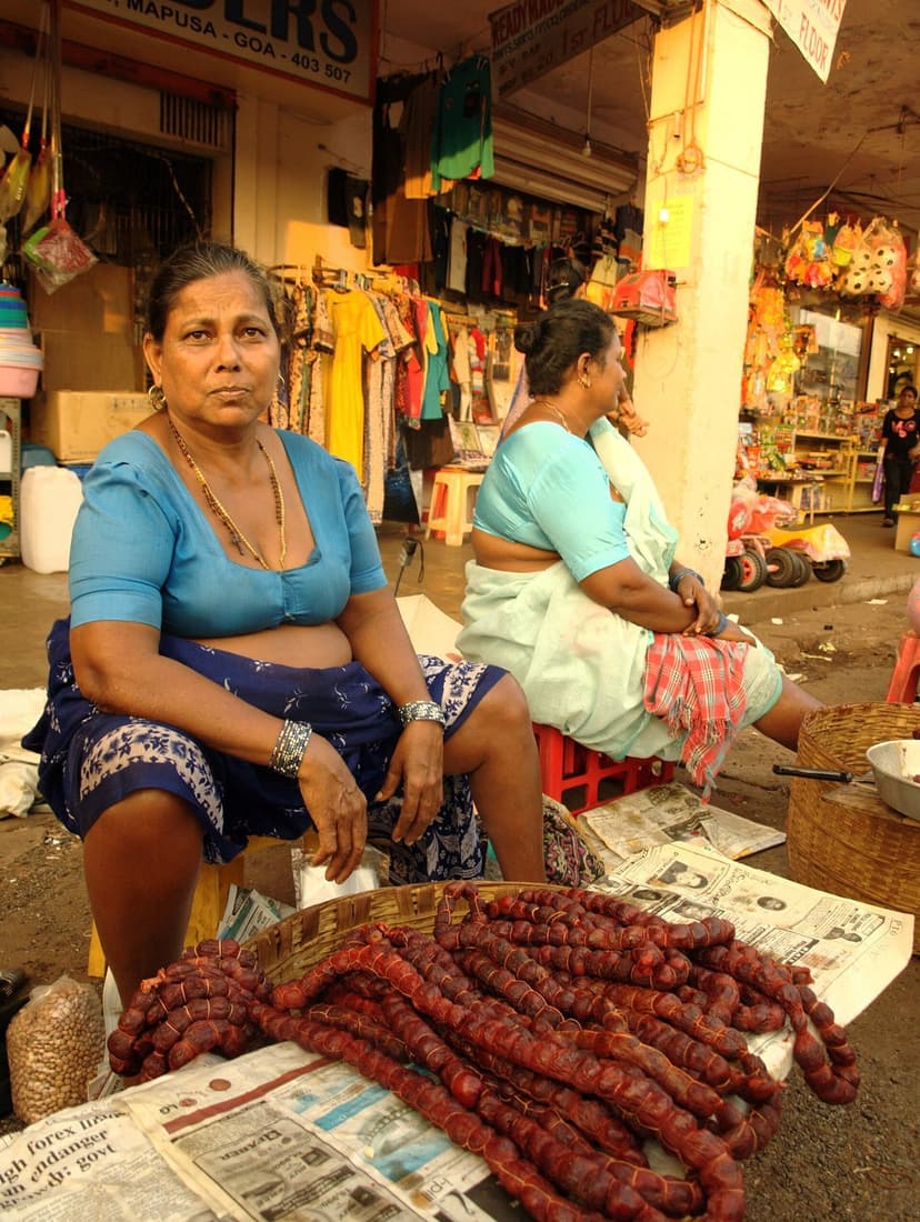 A woman selling strings of dark red Goan chourico sausages laid out on newspaper at the Mapusa market, shops behind and a Mapusa Goa sign visible