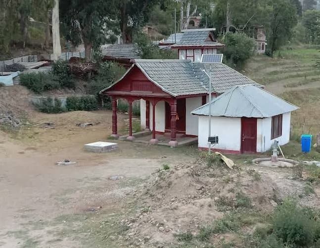 Small Himachali village shrine with red wooden porch and slate roofs in Karsog Mandi, terraced fields and pine ridge in the background