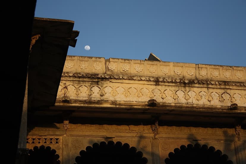 The carved cream stone eaves and cornice of Bagore Ki Haveli in Udaipur with a rising moon above and arched windows below in evening light