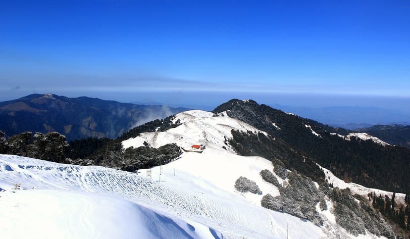 Shikari Devi Wildlife Sanctuary ridge covered in snow, with a small red-roofed structure on the crest against a clear blue sky and Himalayan haze layers below