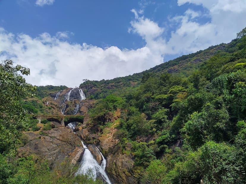 The four-tiered Dudhsagar Falls in Goa in full flow down a green forested mountainside, with an old stone railway bridge crossing in front of the cascade under a blue sky.