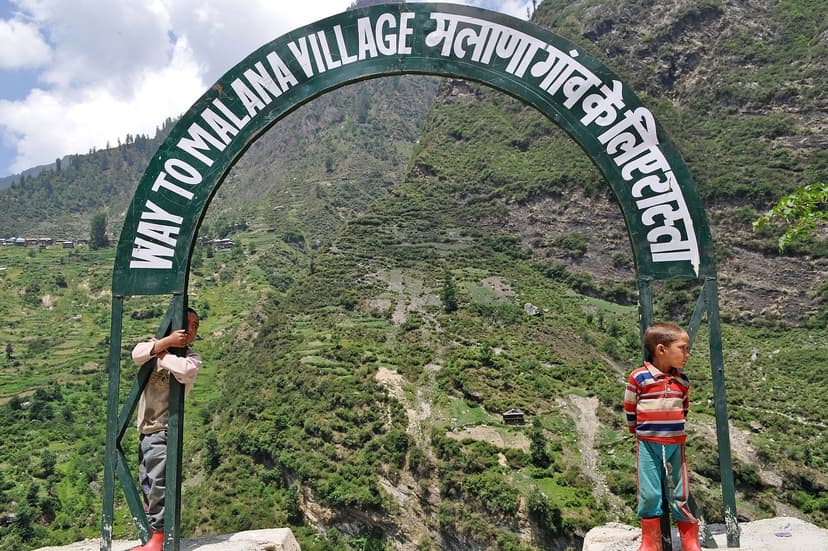 Green metal gateway sign reading Way to Malana Village in English and Hindi, two local children standing beside it, terraced fields behind