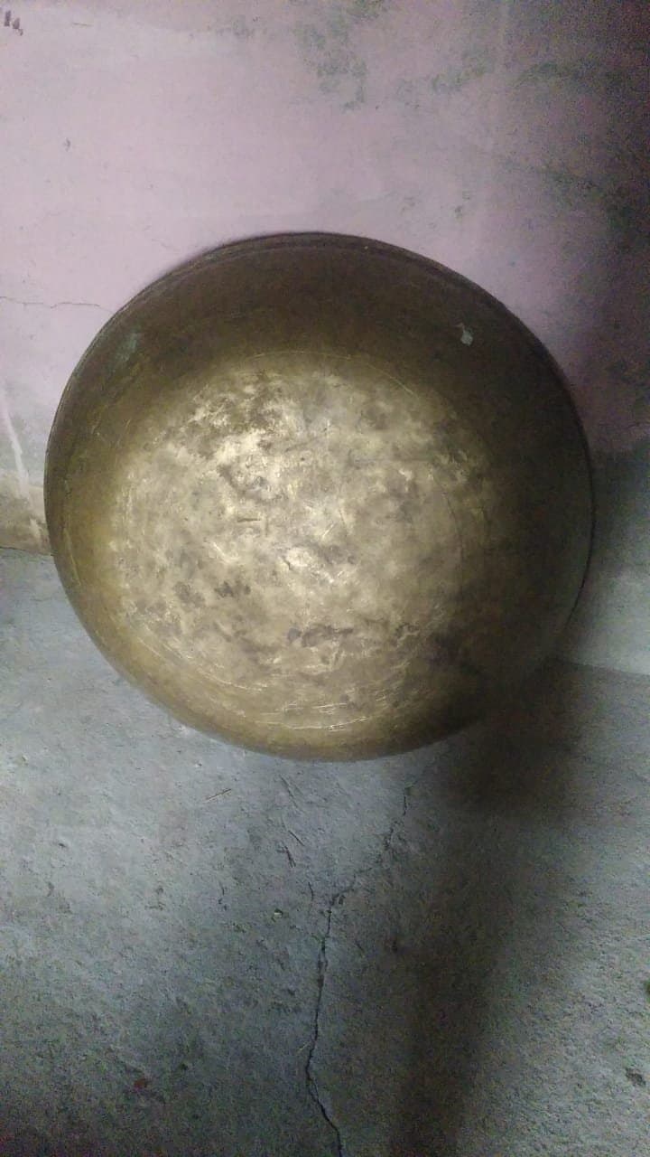 A heavy old brass paraat propped against a stone wall in a village house in Himachal Pradesh — the kind of darkened, well-used brass vessel that Boti families pass between neighbours for community feasts