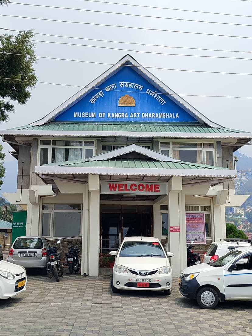 The Museum of Kangra Art building in Dharamshala with its blue-and-yellow signboard in Hindi and English over a covered porch and parked cars at the entrance