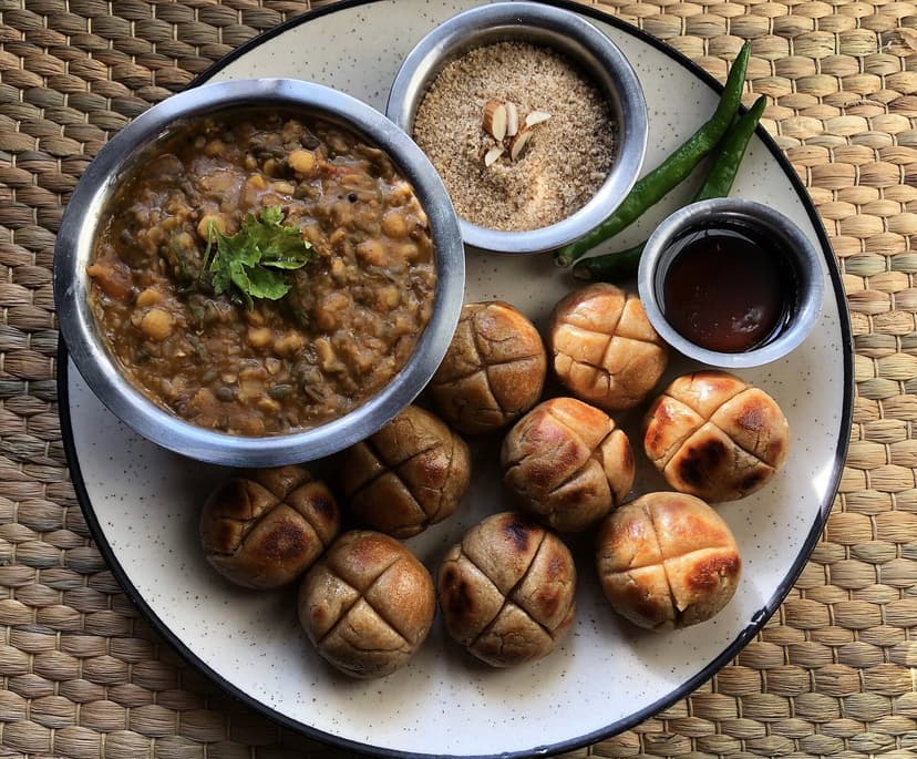 A plate of dal baati churma, baked wheat baati balls with a bowl of dal, a mound of sweet churma, a green chilli and tamarind chutney, on a woven mat