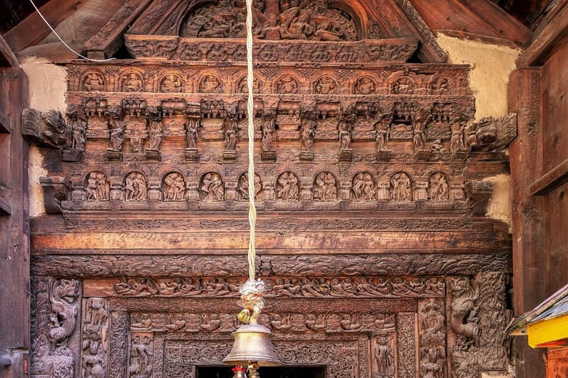 Close-up of the carved wooden lintel of the Lakshana Devi Temple — multiple registers of figures, deities and arched niches in dark wood, with a brass devotional bell hanging in the centre