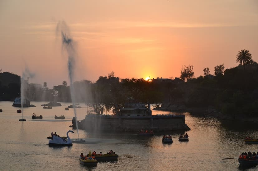 Sunset over Nakki Lake in Mount Abu, Rajasthan, with pedal boats and fountains on the water and silhouetted hills under an orange sky.
